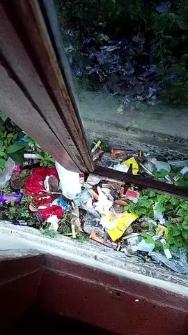 The video shows a pile of garbage in what appears to be an outdoor environment. The garbage consists of various items including plastic bags, food wrappers, fabric, and other debris. There are also some green plants growing amongst the trash. A metal bar or beam runs diagonally across the frame, obscuring part of the scene. The lighting suggests it may be nighttime or a dimly lit area.