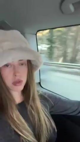 A young woman is in the backseat of a car. She is wearing a light gray, plush bucket hat and a dark gray, long-sleeved shirt. She has long, light brown hair. She is looking directly at the camera with a somewhat pouty expression. Trees can be seen through the car window, indicating that the car is moving. The interior of the car is visible, including the ceiling and a grab handle.  The video appears to be filmed by the woman herself, likely using a smartphone.