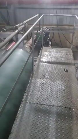 The video shows industrial equipment from a medium height perspective. Two red shafts with universal joints are visible, connecting to motors and machinery. The equipment is located on an elevated platform with a diamond-patterned metal surface and a gray metal railing. Steam or vapor is rising from one of the large cylindrical structures. The surroundings appear to be an industrial building, with pipes, overhead lighting, and metal structural elements visible.