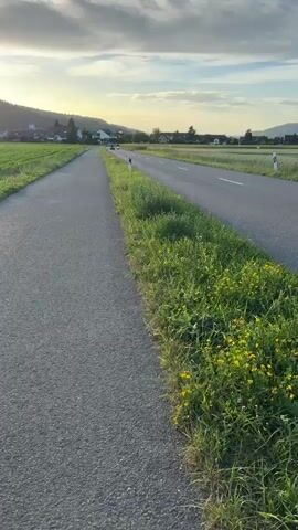 The video begins with a stationary, low-angle shot of a paved road with a narrower paved path to its left. Lush green grass and yellow wildflowers line the right side of the road, separating it from a field. In the distance, the road curves slightly as it passes through a rural landscape with fields and scattered buildings, including a church steeple. The sky above is partly cloudy with a warm, golden hue, suggesting either sunrise or sunset. As the video progresses, two ATVs are seen approaching from the distance on the road. The first ATV, a dark colored quad bike, is ridden by a person wearing dark clothing and a helmet. The second ATV, also a dark colored quad bike, follows closely behind. Both vehicles move along the road at a moderate speed. The camera remains stationary throughout the clip, focusing on the road and the approaching ATVs. The video ends as the two ATVs pass the camera.