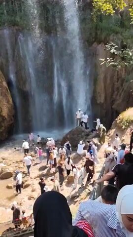 A large crowd of people, dressed in a variety of clothing including many in traditional religious attire, are gathered around the base of a waterfall. The waterfall cascades down a rocky cliff face into a pool of water. Many people are standing in the water, some are walking on the rocks, and others are observing from a raised walkway with a railing. The area is lush with green foliage and trees. The sun is shining, casting shadows on the scene.