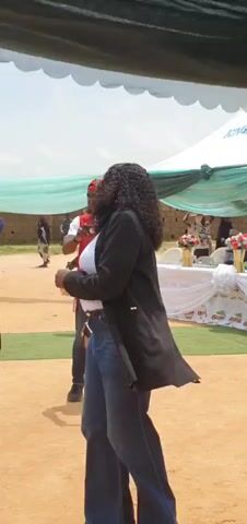 A woman with curly dark hair and sunglasses walks in front of the camera, wearing a white top, a black blazer, and blue jeans. Behind her, a man in a red vest and a red cap holds a phone, seemingly filming. To the left of the woman, another man claps. In the background, there is a tent with green and white fabric, tables with decorations, and other people. A man on the far left is wearing a blue shirt and dark pants. To the left of the woman and in the background, a man is playing a drum. The ground is covered in sand.