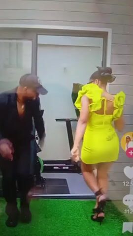 A man in a black outfit and a cap is dancing with a woman in a neon yellow dress and black heels. They are on a green turf area in front of a glass door. Behind the door, there is exercise equipment, including a stationary bike. The woman is animated and expressive, while the man is also moving to the music. A plant is visible in the background.