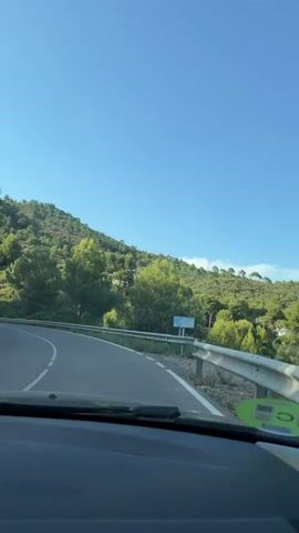 The video shows a view from the inside of a car driving on a winding road. The road is paved and has white lane markings. A metal guardrail runs along the right side of the road. The landscape is hilly and covered with dense green trees and vegetation. The sky is clear and blue. There is a blue sign visible on the right side of the road, partially obscured by trees. A green sticker with a black symbol is affixed to the car's dashboard in the foreground. The sunlight suggests it is daytime.