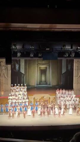 A stage full of performers is shown in a full shot. The backdrop is a projected image of an Egyptian temple corridor. The group of performers is arranged in rows, each wearing different costumes indicative of ancient Egyptian attire. Some are in white robes, others in blue, and some are bare-chested in tan. A man in a dark suit walks towards the group. The theater is darkened with spotlights shining on the stage, and applause can be heard.