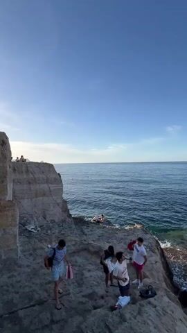 A full outdoor scene is captured from a high-angle view, depicting a group of people at the beach on a sunny day. The camera pans from left to right, revealing a clear sky and the vast expanse of the ocean. On the left side of the frame, a tall, rugged rock formation stands, while a group of people is visible on the beach. Some individuals are seen swimming in the water, while others are relaxing on the shore.