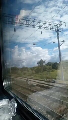 The video captures a view from a train window looking out at a rural landscape. Train tracks run parallel to the train. The sky is blue with fluffy white clouds. The surrounding area is green with trees and foliage. There are overhead power lines for the train. The window has some reflections and what appears to be a plastic bag on the interior ledge.