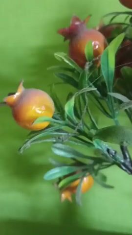 A close-up video shows a fake pomegranate tree with bright red and orange fruits and green leaves. The tree is in a white rectangular pot. The background is a bright green wall. The camera slowly zooms out revealing more of the tree and the white pot sitting on a shelf against the green wall. The video is about 5 seconds long and is shot from a side angle.