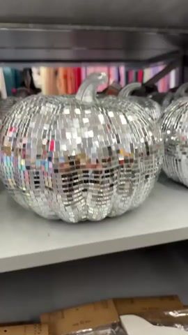 The video shows a close-up shot of a silver disco ball pumpkin. The pumpkin is covered in small, square mirrored tiles that reflect light. It has a clear, glass-like stem. The pumpkin is sitting on a white shelf, and other disco ball pumpkins are visible in the background. The shelf appears to be in a store, as some items are displayed on shelves below. The overall impression is of a festive and sparkly decoration.