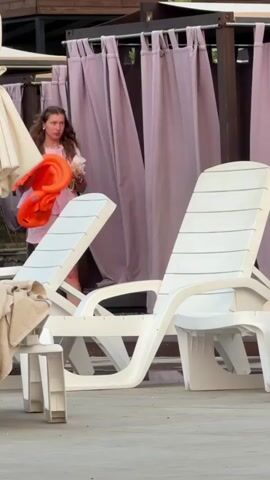 A woman in a pink outfit walks towards the right holding an orange inflatable object and a white object. Another woman in a black garment carries a baby. The setting appears to be a poolside with lounge chairs and cabanas.