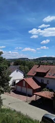 The video captures a scenic view of a suburban neighborhood on a clear, sunny day. The sky is a vibrant blue, dotted with fluffy white clouds. In the foreground, there are several residential buildings with red-tiled roofs and white or light-colored walls. One building has a prominent white facade and a smaller attached red section with garage doors. Another larger building features multiple windows and a dark-colored chimney. A covered carport with a tiled roof is also visible. Trees with green foliage surround the houses, and a well-maintained lawn is visible in the background. Further in the distance, rolling hills covered in trees and scattered houses form a picturesque landscape. The overall impression is one of a peaceful and idyllic residential area bathed in bright daylight.