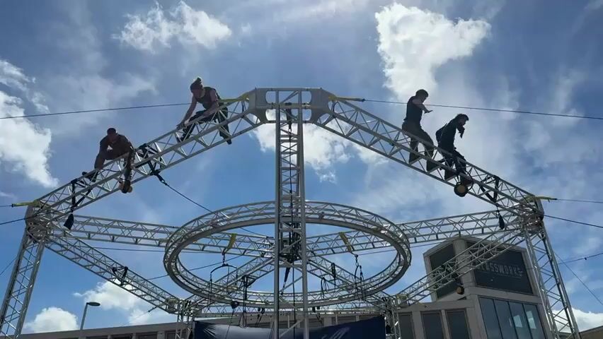 Four people are positioned on top of a large metal structure against a sky with scattered clouds. The structure is a complex arrangement of trusses and circular elements, resembling a stage or performance platform. The people appear to be setting up or performing, moving around on the structure. The scene is outdoors, and the building "Glassworks" is visible in the background. The video is shot from a low angle, emphasizing the height and complexity of the structure.