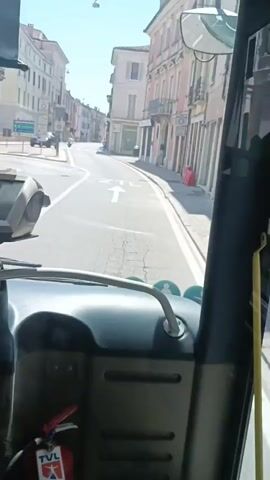 The video is filmed from the interior of a bus looking out the front windshield. A fire extinguisher is visible on the lower left inside the bus. The bus is driving down a narrow street lined with multi-story buildings, likely in a European town. The buildings have architectural details such as balconies and shuttered windows, and many appear to be shops or businesses on the ground floor. There are signs on the buildings and on poles along the street. A white arrow painted on the road indicates a lane for vehicles. A white sign with a circular red border, indicating a restricted zone, is visible. Two people on scooters are seen further down the street. A pedestrian walks on the sidewalk. The sky is clear and blue, indicating a sunny day.