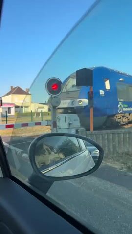 A train passes by a car stopped at a railway crossing. The train is blue and white with green accents and displays the text "Région Hauts-de-France". The car's interior is visible, including the steering wheel and dashboard. A person's hand is resting on the open car door. The sky is clear and blue.