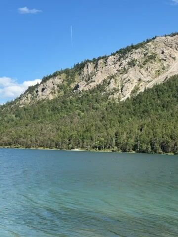 The video shows a serene lake with a mountain backdrop. The water is a clear turquoise, with gentle ripples on its surface. Dense green trees cover the lower slopes of the mountain, giving way to rocky, barren terrain higher up. The sky is a vibrant blue with a few wispy white clouds and a thin white contrail from an aircraft. The sunlight appears to be bright, casting a pleasant glow on the scene. There are no people or man-made objects visible in the majority of the frames, creating a sense of natural tranquility. In the last few frames, a dark object, possibly a sign or a piece of equipment, briefly enters the bottom left corner of the frame. The camera appears to be moving slightly, perhaps from a boat or a stable elevated position on the shore.