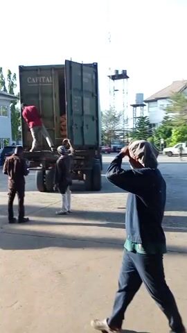 A video shows a shipping container being unloaded by workers. The container is labeled "CAPITAL".  A man in a red shirt is visible inside the container. The camera follows a man walking toward a large metal door. The door opens, revealing the interior of a warehouse filled with stacks of boxes, black pallets, and blue pallets.
