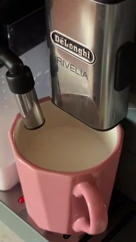 In this close-up, medium shot, a DeLonghi Rivelia coffee machine is in operation, dispensing a beverage into a pink, faceted mug. The machine, primarily silver and black, has the "DeLonghi" and "RIVELIA" logos clearly visible on its stainless steel exterior. A thin stream of dark brown liquid, presumably coffee or espresso, pours from the machine's nozzle into the mug, which already contains a frothy, white liquid, likely steamed milk. The mug has a prominent handle and is filled to about two-thirds capacity. The scene appears to be indoors, with muted lighting, suggesting it could be any time of day. The setting is domestic or commercial, likely a kitchen or café, given the presence of the coffee machine and mug. There are no people visible, and the focus is entirely on the coffee-making process. The mood is quiet and focused, highlighting the simple act of preparing a coffee drink.