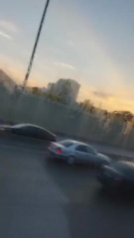 A video taken from a moving vehicle shows a highway with cars driving on it. In the background, there are buildings and trees. The sky is light blue with some clouds. The sun appears to be setting.