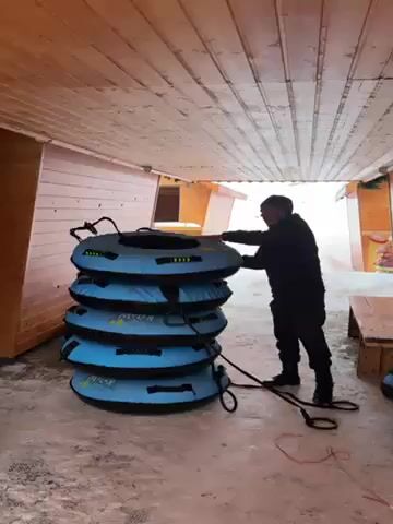 A man in black clothing and a hat is taking a blue inflatable snow tube from a stack of similar tubes. The tubes are stacked in a covered area with a wood ceiling and walls, suggesting a rental or storage space near a snowy area. There is a wooden bench visible on the right side. The ground is covered in a mix of snow and possibly dirt. A rope is attached to the bottom tube.