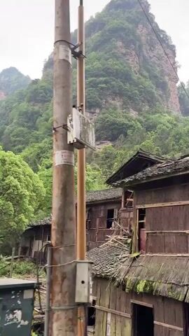 The video shows a picturesque landscape in a mountainous region. An old, weathered wooden structure with a decaying roof is in the foreground. The building seems to be in disrepair, with moss and discoloration on its walls. Behind it, a towering mountain covered in lush greenery dominates the background. The camera then pans to show a parking area with several vehicles parked under a covered walkway. The walkway has a traditional Asian design, which complements the scenic backdrop of the mountains. A person stands near a white car, looking towards the camera. A few young trees are planted within the parking area, and the overall setting appears to be a tourist spot or a site of natural beauty.