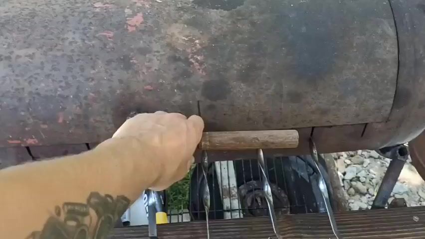 The video shows meat skewers cooking over charcoal in a makeshift grill. A hand with tattoos is visible briefly at the beginning of the video, opening the grill lid. The grill appears to be made from a metal barrel cut in half lengthwise, with skewers resting on supports along the top edge. The meat is browned and appears to be chicken or pork. The environment is outdoors, with greenery visible in the background. The video appears to be a close-up view of the cooking process, with no other people visible.