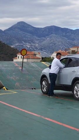 The video shows a man standing next to a silver Nissan Qashqai on what appears to be a ferry or similar vessel, with a mountainous landscape in the background. The man is wearing a white long-sleeved shirt and jeans, and he is leaning against the driver's side door of the car. A sign with a red circle and a horizontal white bar is planted behind the car near a ramp that leads up. The floor of the ferry is painted green with orange lines marking lanes. A second man walks into the frame, coming from behind the car towards the left. He's wearing a dark blue jacket with "JP MD" in orange letters on the back.