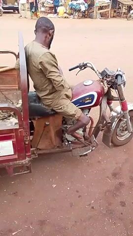 A man in a tan outfit sits on a burgundy motorcycle-rickshaw, with his back to the camera, appearing motionless. The rickshaw is parked on a reddish-brown dirt surface in what looks like a marketplace. Other motorcycles and people are moving in the background, suggesting a bustling environment. The rickshaw has a small flatbed cargo area behind the motorcycle. The rickshaw's cargo bed is empty except for some loose items. The video captures the scene in a stationary shot, offering a view of the immediate surroundings and the activity within the marketplace setting.