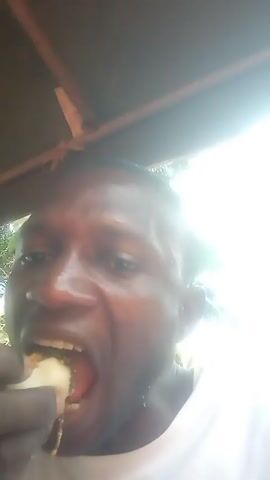 A man eats while filming himself in close-up. He is wearing a white t-shirt, and appears to be seated under a wooden structure. He seems to be enjoying his food, but it's difficult to tell what he's eating. The background is mostly bright due to sunlight.