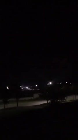 The video shows a dark, outdoor scene at night. Several streetlights are visible in the distance, illuminating a paved area that appears to be a road or parking lot. Silhouettes of trees are present in the foreground and midground. The overall atmosphere is quiet and still.