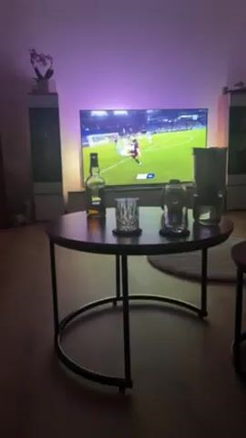 The video shows a living room setting with a round coffee table in the foreground. On the table are a bottle of liquor, a cut glass, and a glass teapot, all on coasters. Behind the table is a large television displaying a soccer game. Players are in action on the field, and the goalkeeper, wearing a bright yellow jersey, is making a save. The room is lit with soft, purple-toned ambient light, possibly from LEDs behind the TV, which casts a glow on the wall. Flanking the TV are tall speakers. A potted orchid is visible on top of one of the speakers. The room appears tidy and modern.