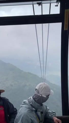 The video shows the interior of a cable car with people inside. Outside the window, a mountainous landscape shrouded in mist is visible. Several steel cables extend upwards, disappearing into the sky. In the distance, a few red cable car cabins can be seen suspended on the cables, moving along the route. On the left side of the cable car, a woman wearing a straw hat and a red shirt with a dragonfly print is seated. Next to her, a person wearing a white baseball cap and a light-colored jacket, with a face mask covering their lower face, is also seated. A younger person in a blue shirt is visible on the far left. A red and white sign with QR codes is affixed to the interior wall of the cable car. The overall impression is of a scenic journey through a misty mountain range via a cable car.