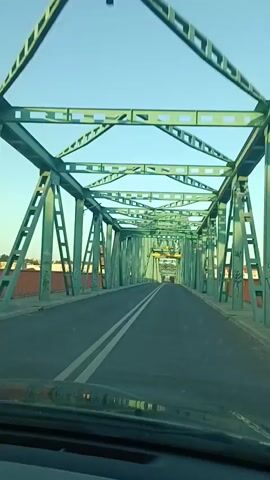 The video begins with a first-person perspective from a vehicle driving across a green metal truss bridge. The bridge has a two-lane asphalt road with white dividing lines. On either side of the road are orange railings. The camera pans to the right, revealing a body of water below the bridge, which appears to be a river or a large canal. The water is a greenish-blue color, and there are gentle ripples on its surface. The sky is a clear, pale blue, suggesting it is either early morning or late afternoon. The far bank of the waterway is lined with trees and some structures are visible in the distance. The video continues to show the view from the car as it drives across the bridge, with the water and distant landscape remaining visible to the right. The perspective then shifts slightly to show the view through the car's side window, again focusing on the water and the bridge's railings. The final shots continue to show the water and the landscape, with the bridge structure becoming less prominent. No people are visible in the video, and the actions consist solely of the vehicle driving across the bridge.