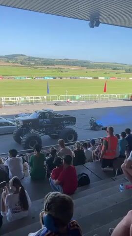 The video captures an outdoor event with spectators seated on tiered bleachers facing a flat track. The track itself appears to be asphalt with a grassy field and hills in the background. Several large monster trucks are present on the track, some with elaborate designs and massive tires. In the foreground, people of various ages are watching the event, with some holding cell phones. The lighting suggests it is daytime with clear skies. The actions include a monster truck positioned near the center of the track, and another monster truck, red in color, is also visible. There is also a smaller, dark-colored vehicle on the track. Blue and pink smoke are being emitted from some of the vehicles.