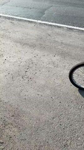 A person in camouflage clothing rides a red bicycle with a basket on the front and a rack on the back down a paved road. The person is riding on the shoulder of the road, which is made of gravel and dirt. A large semi-trailer truck passes the cyclist on the road. The sky is cloudy and the sun is shining. There are trees and fields on either side of the road. The cyclist is moving away from the camera.