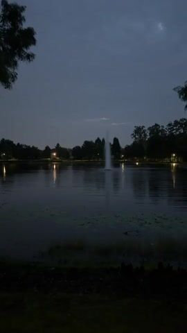 The video shows a lake at dusk or dawn. A fountain shoots water into the air in the center of the lake. Trees and lights are visible on the far shore. The sky is cloudy and dark.