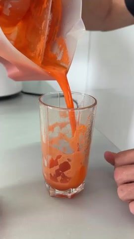 A close-up shot captures a person's hand pouring bright orange juice from a blender cup into a clear, ribbed glass. The juice cascades into the glass, creating a frothy layer as it fills. The glass sits on a light-colored countertop, and a portion of a white appliance is visible in the background. The scene appears to be in a kitchen setting.