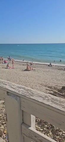 The video shows a crowded beach with many people sunbathing and swimming. The beach is made of pebbles, and the water is a clear blue. The sky is also clear and blue. There is a wooden fence in the foreground, and some debris is visible on the beach. The video pans across the beach, showing different groups of people. Some people are lying on towels, while others are playing in the water. There are also a few boats in the distance. The overall impression is one of a lively and enjoyable beach scene.