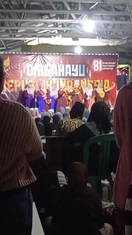 The video shows an indoor event with a stage at the back. On the stage, there are approximately ten women standing in a line, wearing matching purple dresses and sashes. They appear to be holding microphones and are facing a crowd of people. A banner behind them reads "DIRGAHAYU REPUBLIK INDONESIA" with the number "81" prominently displayed. The audience members are seated in chairs and on the floor, facing the stage. The lighting is dim, with some overhead lights illuminating the stage. The environment appears to be a community hall or a covered outdoor area. The video is shaky, suggesting it was recorded handheld.