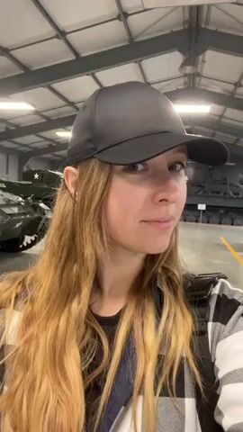 A woman with long, blonde hair and a black baseball cap is seen in a selfie-style video. She is wearing a black and white plaid shirt over a black t-shirt. The background appears to be a large, industrial-style building with a high ceiling and exposed beams. In the blurry background, several large military vehicles, resembling tanks, are visible. The lighting is bright, suggesting artificial lighting within the building. The woman is looking towards the camera and slightly smiling. The video is short, capturing a brief moment of her looking around.