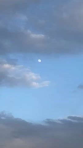 The video opens with a close-up shot of the full moon set against a clear blue sky, partially covered by scattered white and grey clouds. The camera slowly pans downwards, revealing more of the sky and the lower cloud formations. As the pan continues, a vast, arid landscape comes into view, characterized by rolling hills covered in dry, light brown and green brush or grass. In the mid-ground, a tall utility pole with what appears to be multiple lines stretches towards the sky from the top of a distant hill. The sky remains mostly blue with clouds throughout the pan. The scene suggests a daytime setting, possibly early morning or late afternoon, given the visibility of the moon. The overall impression is one of a serene, open, and rural environment. No people or visible text are present.