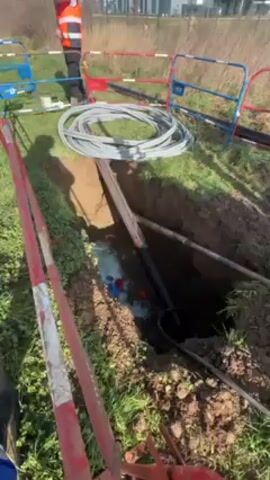 A medium-shot video shows a construction site with a trench dug in a grassy area, bordered by red and white and blue and white safety barriers. A construction worker in an orange vest stands in the background. Inside the trench, water has collected, revealing pipes of various colors (red, blue, black). A large black pipe with a red stripe runs along one side of the trench. A coil of gray tubing rests on the grass next to the trench. The camera perspective is from ground level, looking down into the trench. The ground around the trench is dirt and grass. The overall scene suggests ongoing or recently completed construction work involving underground utilities.