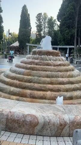 A stepped fountain with water flowing down its tiers is prominently featured. The fountain is made of stone with bands of different colors and textures. In the background, trees and a clear sky are visible. Several people are present in the scene, including a person pushing a stroller, two individuals walking, and a few others in the distance. The ground around the fountain appears to be paved. The overall environment suggests an outdoor public space, possibly a park or plaza, during the daytime.
