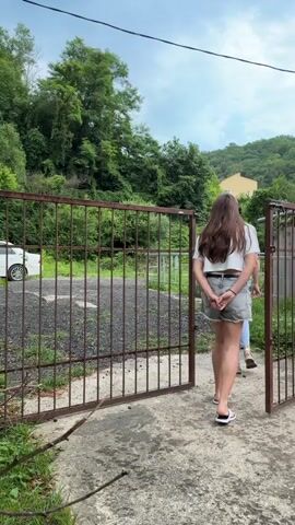 A young woman with long brown hair and wearing a white t-shirt and gray shorts is standing near an open metal gate. She has her hands clasped behind her back. Another person, partially visible, is standing behind her. The gate is made of vertical metal bars and appears to be rusted. The ground inside the gate is covered with gravel. Beyond the gate, there is a white car parked. The background consists of lush green trees and vegetation, with a cloudy sky overhead. A yellow building is visible in the distance on a hillside. The scene appears to be outdoors in a somewhat overgrown area. The woman then turns and appears to be interacting with the person behind her.