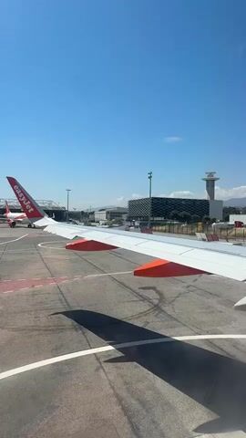 This is a video taken from the window of an airplane, showing the wing of the plane in the foreground. The wing is white with orange accents on the underside. The plane is on the tarmac of an airport. In the background, there is another airplane, an airport building with a modern facade, and an air traffic control tower. The sky is clear and blue. The overall scene suggests the plane is either about to take off or has just landed.