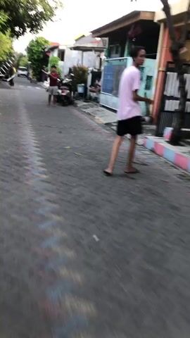 The video shows a street scene in Kalisampurno, Indonesia. The time of day appears to be late afternoon or early evening, given the soft lighting and long shadows. The street is paved with dark cobblestones, and there are houses lining both sides of the street.

In the foreground, a person wearing a pink shirt and black shorts is walking away from the camera. In the middle ground, a man in a red shirt and shorts is standing near a parked motorcycle. Further down the street, there is a pile of dark gravel and some construction materials, including white blocks. Several men are working around the gravel pile. One man is holding a long ladder, appearing to be preparing to move it or set it up. Another man in a yellow shirt is near the gravel. The overall atmosphere seems to be one of ongoing work or preparation for construction.

There are no visible signs with text in this clip. The weather appears to be clear. The scene depicts a typical residential street in Indonesia with some construction activity taking place.