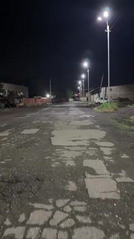 The media file captures an outdoor scene at night in Argavand, Armenia. The primary subject is a damaged asphalt road, characterized by numerous cracks and patches, suggesting wear and tear. The road is illuminated by a series of modern streetlights mounted on tall poles, casting a bright, artificial glow.

The setting appears to be a semi-industrial or less developed area. On the left side of the road, there are piles of what seem to be scrap metal or discarded materials, with a low-rise building or structure behind them. Further down the road, on the same side, a red light is visible, possibly from a vehicle or a traffic signal.

On the right side of the road, there are more structures, including what looks like a white utility vehicle or van parked near the curb, and a brick building further in the background. There is also some vegetation, mostly dry grass, lining the edge of the road. The sky is dark and clear, indicating nighttime and likely fair weather.

No people are visible in the media file. The overall impression is one of a quiet, utilitarian area, possibly on the outskirts of a town or city, with infrastructure in need of maintenance. The activity taking place is simply the illumination of the scene by the streetlights. The time of day is clearly night. There is no visible text in the media file.