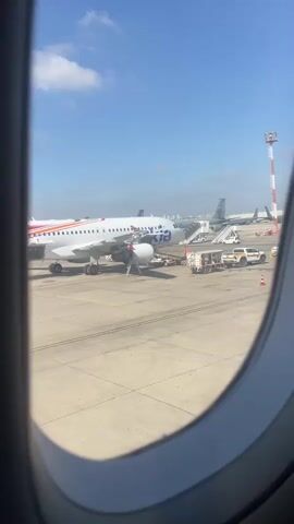 The video is shot from inside an airplane window looking out at an airport tarmac. A white airplane with red and blue accents is parked at a gate. A set of stairs is connected to the plane, and several airport vehicles, including a small white truck, are present. In the background, other airplanes and airport infrastructure are visible under a clear blue sky with scattered clouds. The video is short and static, offering a glimpse of the airport environment before departure.