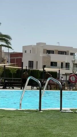 The video shows a residential swimming pool area on a sunny day. In the foreground, the edge of a turquoise swimming pool is visible, with metal handrails leading into it. The area around the pool is covered in artificial green turf. In the middle ground, several people are present. One person, wearing a dark swimsuit, is seen jumping into the pool, causing a splash. Another person, also in a dark swimsuit, is in the pool. Further back, other individuals are sitting or walking around. A red life preserver is mounted on a fence. In the background, there is a modern, multi-story white building with balconies and windows. Various trees, fencing, and tents are also visible. The sky is clear and blue.