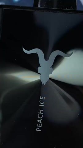 The media file is a close-up, static shot of a black surface, likely a product or electronic device. A stylized grey silhouette of a goat's skull with large, curved horns is centered on the surface. Below the skull, the text "PEACH ICE" is printed vertically in a grey, sans-serif font. The surface has a reflective, brushed metal texture, causing light to refract and create streaks of bright white and deep shadow radiating from the center. The overall impression is sleek and modern, with a slightly edgy or symbolic aesthetic due to the goat skull imagery. There are no people, animals, or an obvious setting visible beyond the product itself. The lighting suggests an indoor environment, possibly with artificial light sources causing the reflections.