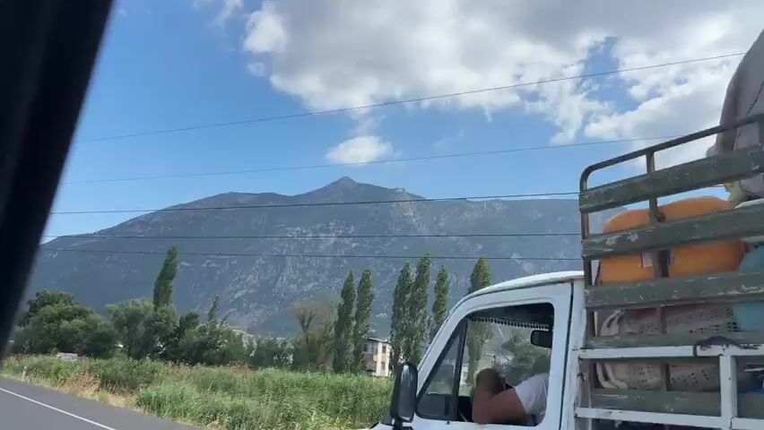 This is a short, low-angle video shot from a moving vehicle, likely a car, looking out a window. The viewpoint is from the passenger side, evidenced by the interior framing and the curve of the window. The video captures a rural landscape with a dominant mountain range in the background. The sky is a bright blue with scattered white clouds. In the middle ground, there are several tall, slender trees that appear to be cypress or poplars, interspersed with lower-lying green bushes and dry grass. To the right, there are a few low-rise buildings, some appearing to be residential. A road is visible in the lower left corner of the frame, with the vehicle's tire marking the edge of the pavement. There are power lines stretching across the upper portion of the image. A small signpost with some writing and graphics is partially visible on the left side, in front of some greenery. The scene is static, with the only motion being that of the vehicle. There are no people or animals visible in the frame.